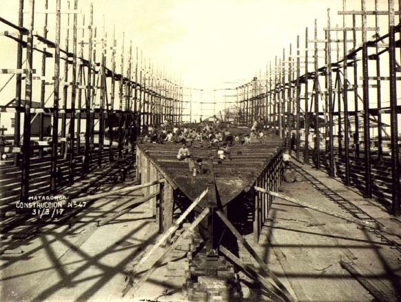 Fotografía histórica de la construcción de un casco de barco entre estructuras de astillero.