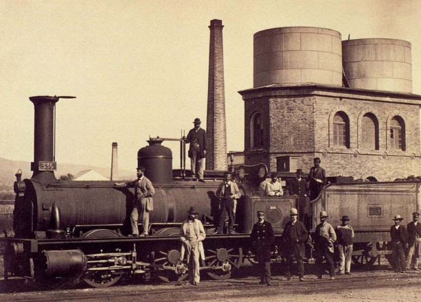 Fotografía histórica de una locomotora y su depósito de agua junto a una estación.